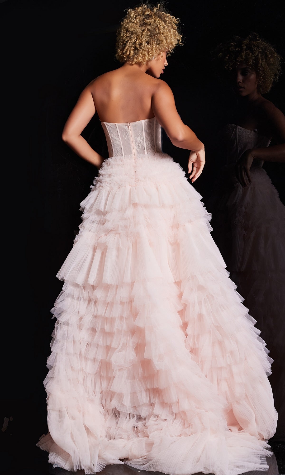A woman with curly hair faces away from the camera in the Jovani Ruffled Long Strapless Prom Ball Gown 38540, light pink with ruffles, as her reflection appears against a black background.